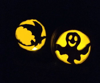 Two carved foam pumpkins glowing with battery tea lights inside in the dark. The one on the right has a happy ghost, and the one on the left is a silhouette of Toothless