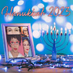 A blue spangled background with a lit menorah and some blue ights on a table, with a framed picture of the Ladies ver 2.0, and in peach letters it says Hanukkah 2023 at the top