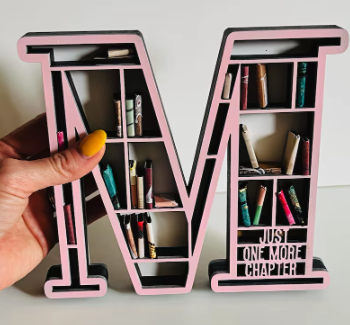 A large pink M with shelves inside, laden with miniature colorful books, and JUNE ONE MORE CHAPTER in the lower corner. It's behind held by a hand with yellow painted nails to show the size 