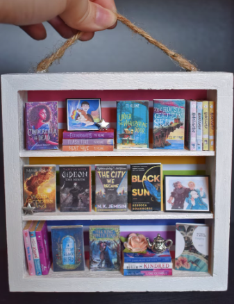 A custom bookshelf with titles like THE CITY WE BECAME, GIDEON THE NINTH, The House in the Cerulean Sea, a tepot and a tiny star 