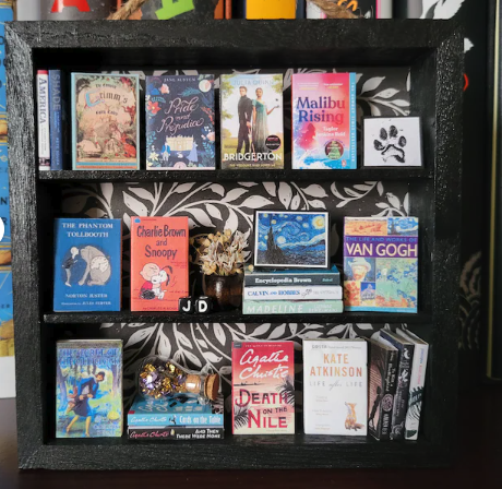A miniature bookshelf with a wallpaper background showing an array of books including The Phantom Tollbooth, Charlie Brown and Snoopy, Pride and Prjudice, Death on the Nile and Bridgerton 