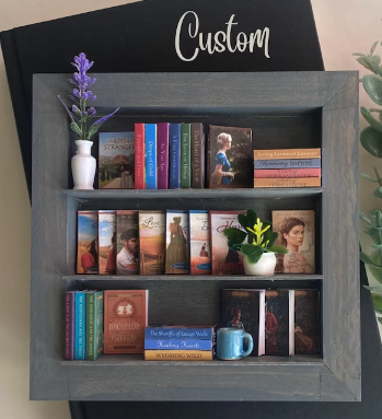 A miniature shadowbox bookshelf with books on each shelf including some inspirational romances and some historical titles like The Bachelor and the Bride, along with a miniature mug and some flowers 