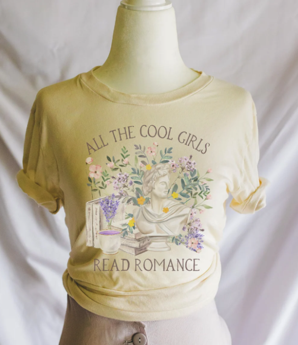 A butter yellow tshirt on a mannequin with a watercolor style illustration of some books, a cup of tea, a bust and some flowers in lilac and pink, with the words All the Cool Girls Read Romance