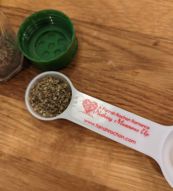 A close up of a measuring spoon with a teaspoon of oregano in it and the handle says A Farrah Rochon Romance - Nothing Else Measures Up