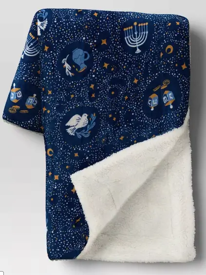 A deep blue fuzzy print with little pictures of menorot, dreidels, doves and oil scattered on a starry background. The reverse side is a plush in white