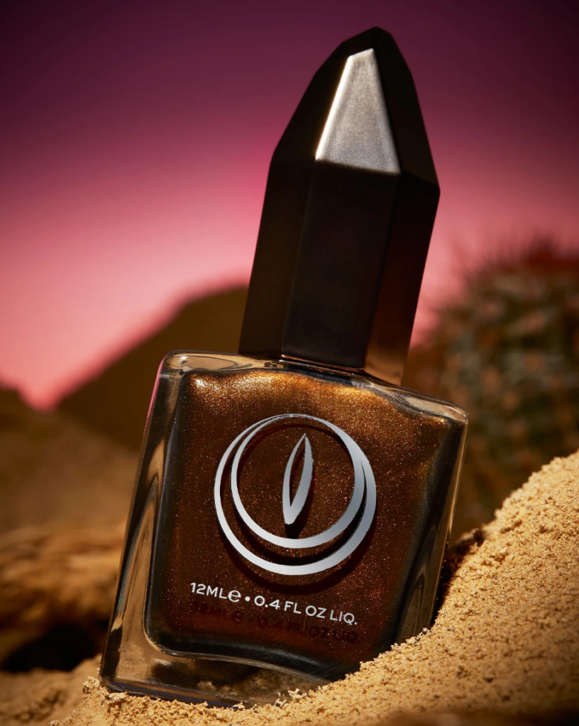 A dark bronze nail polish in a square glass bottle with black cap. The bottle is nestled in sand with a pink sunset background.