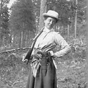 Annie with a dark skirt in a tough material, light colored shirt, and white hat, holding a gun and smiling with a hand on her hip.