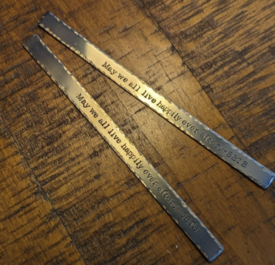 Two bars of bronze with hammered edges against a wood background. The bronze has been stamped to read May we all live happily ever after with a heart, then SBTB