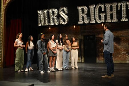 A group of women stand on stage below a sign that says Mrs. Right. Joey stands across from them.