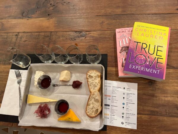 An overhead shot of a tray with four cheese portions with jelly and bread slices. Above the tray are four glasses for wine and a larger glass for water, and a fork. To the right are a copy of The True Love Experiment by christina Lauren and The Lily of Ludgate hill by Mimi Matthews, and below that a tasting guide