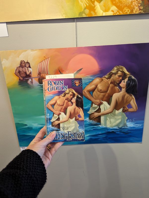 my hand holding the book cover in front of the painting where a blonde fabio dude is shirtless waist deep in water holding a towel maybe around the backside of a white woman with dark hair who is also topless