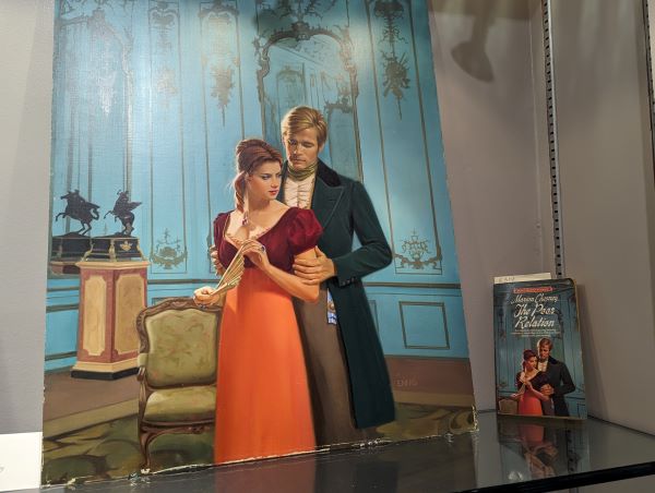 A marion Chesney novel alongside the original art featuring a woman in an orange and burgundy regency gown and a dude in a long coat standing behind her in a blue parlor