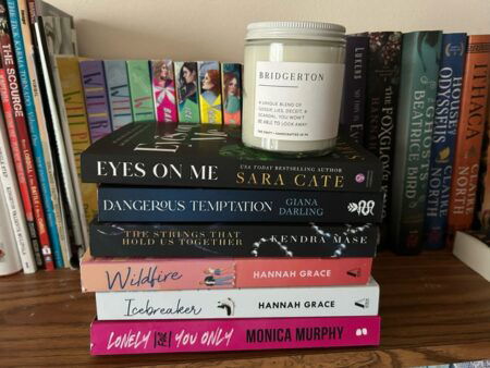 A Bridgerton candle sits on top of a stack of six books: Eyes on Me by Sara Cate, Dangerous Temptation by Giana Darling, The Strings that Hold Us Together by Kendra Mase, Icebreaker and Wildfire by Hannah Grace, and Lonely for You Only by Monica Murphy.