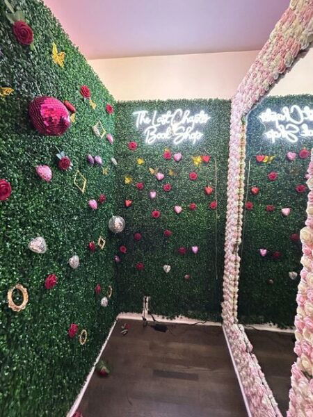 A fitting room with greenery and glittery pink and silver items on the walls. There is a neon sign that reads The Last Chapter Bookshop up high.
