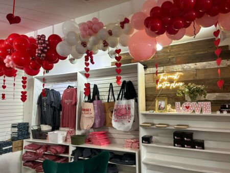 A shelf with bookish tees and tote bags. There are red and pink balloons hanging from the ceiling.