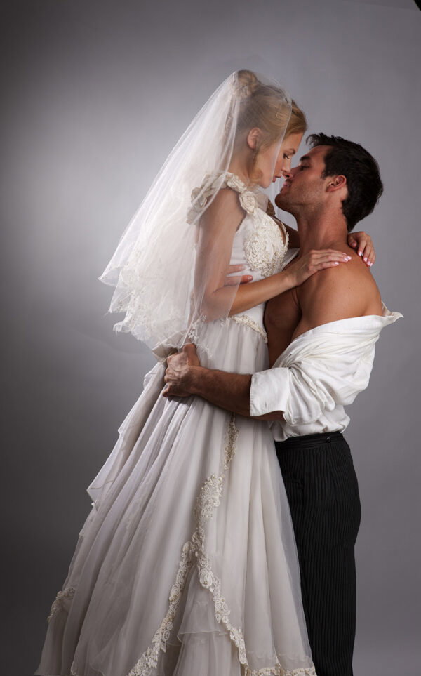A photograph of a white blonde woman in a wedding dress and waist length veil, resting her hand on a male model's chest. His shirt is open but still tucked in OF COURSE and she seems to be standing on a stool 