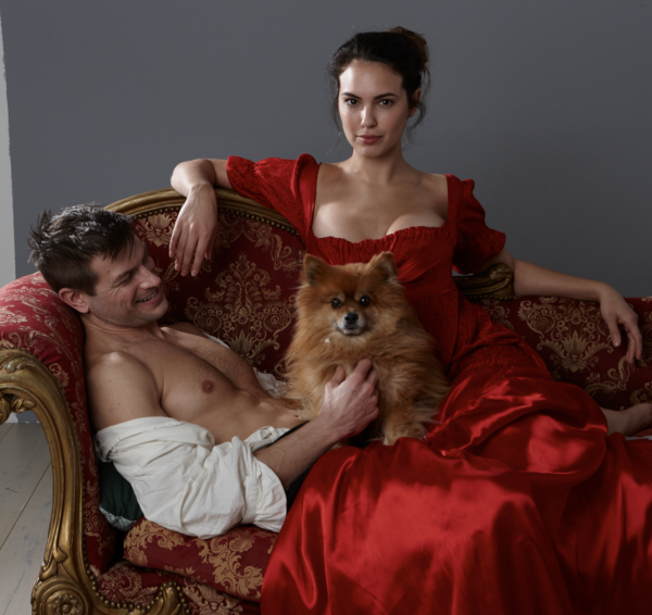 A pair of models recline in mid-pose with Rocky perched on their laps. A woman in a red period gown sits on top of a man laying down in a shirt Unbuttoned But Still Tucked In! And rocky looks very happy. 
