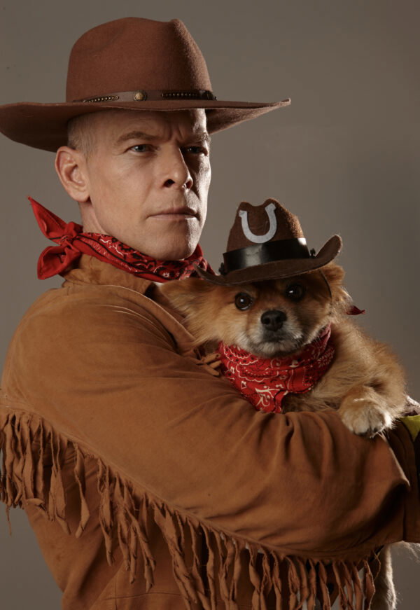 A white man with great cheekbones in a cowboy hat and fringed jacket holds Rocky, who is wearing a bandana and hat to match 