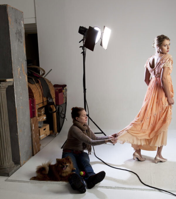 Sharon a white woman with red hair is holding the edge of the model's dress while she looks over her shoulder at the camera. On Sharon's lap is a very happy brown pomeranian and there are props and backdrops visible behind her 