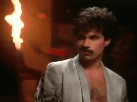 John Oates from Hall & Oates wearing a grey jacket with no shirt, with a flaming torch burning behind him as he looks over his shoulder and smolders