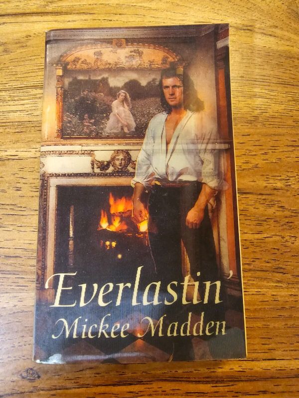 the lenticular cover for Everlastin' which changes based on the angle from which the cover is viewed. The White dude with long hair and a shirt unbuttoned but still tucked in is standing (or disappearing from) in front of a lit fireplace, with a oil portrait of a woman above the mantel