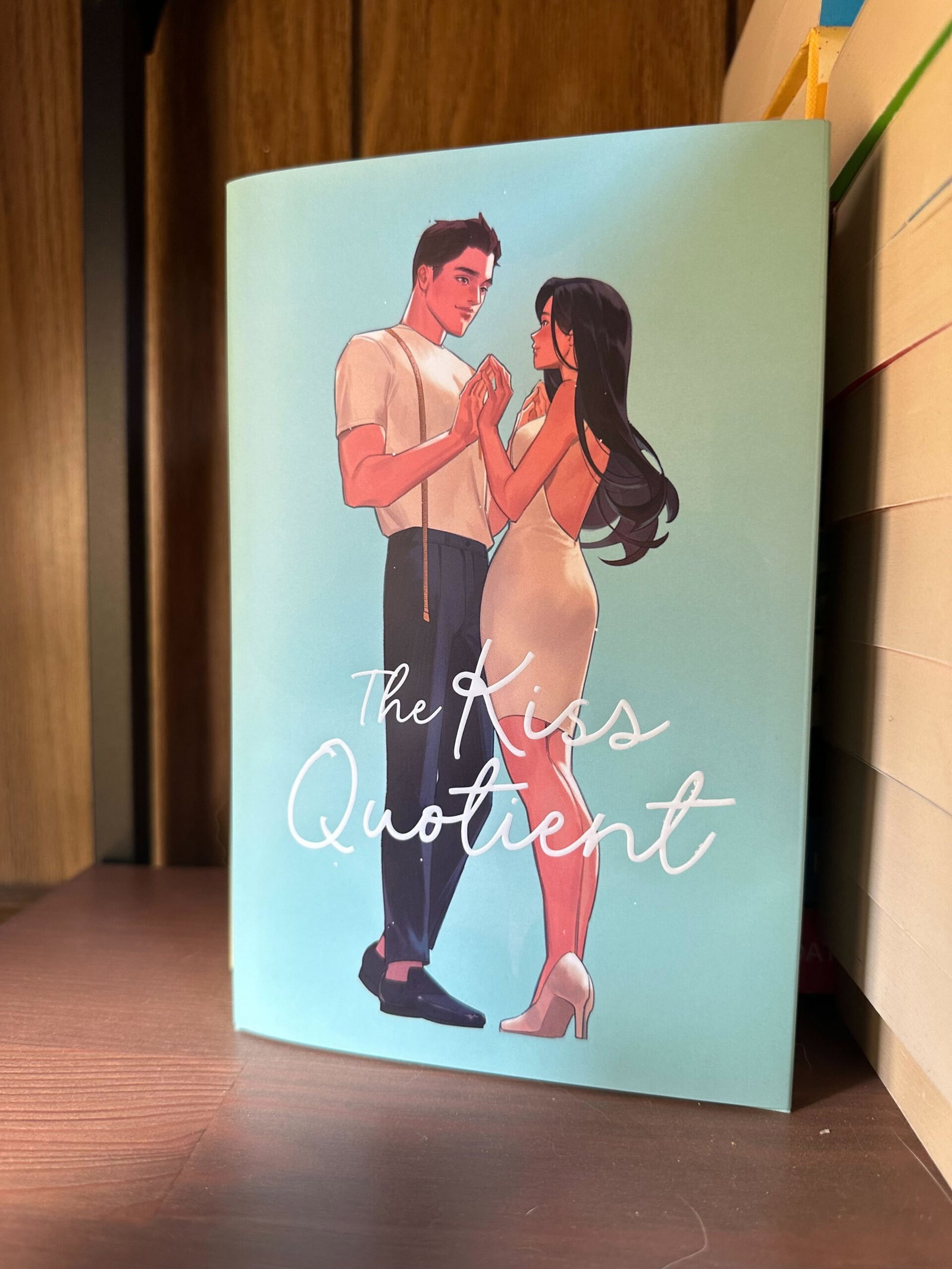 The Kiss Quotient special edition. A pastel blue cover with an illustration of a man in pants and white shirt. He has a tape measure around his neck and he's holding the hands of a woman in a silvery white dress.
