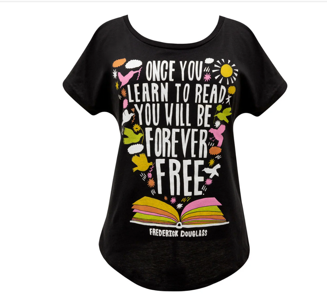 A black shirt with a quote by Frederick Douglass: Once you learn to read, you will be forever be free. The bottom of the shirt has an open book and the quote is surrounded by birds, clouds, and a sun in varying colors of yellow, white, pink, and green.