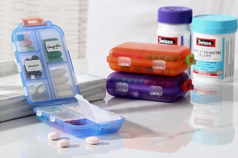 A picture of a folding pillbox on a table with two more pillboxes behind it and some pill bottles. It hinges in the middle so there are five compartments on each side 