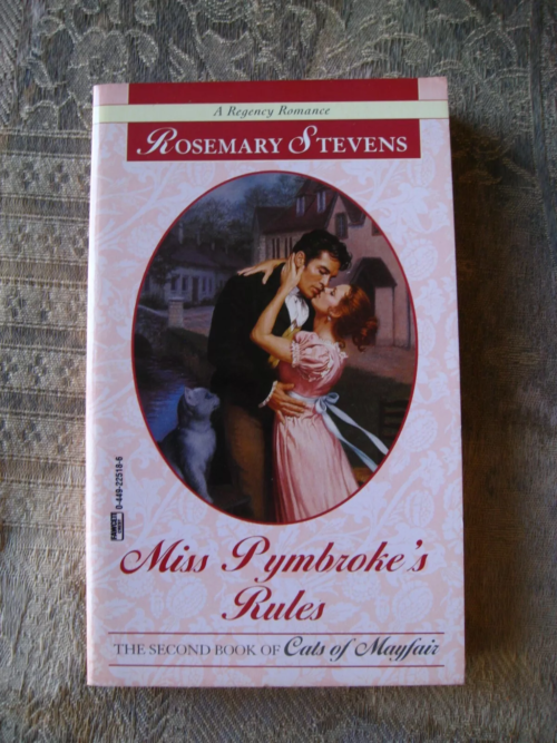 An image from an online listing showing Miss Pymbroke's Rules against a pink lace table cloth. A couple is embracing while a cat stands behind them looking like it is about to sharpen its claws on his backside