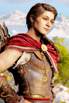 Kassandra standing with her hands on her hips and a red drape over her shoulder she looks bemused
