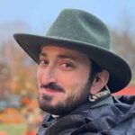 A headshot of Chris DeRosa he is a white man with a mustache and beard wearing a green hat with a brim and a black raincoat, and a producer's earpiece and cord is visible in his ear he is looking over his shoulder and smiling