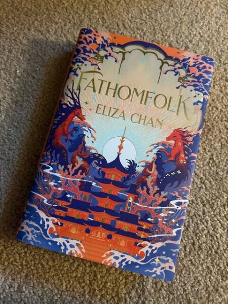 A hardcover copy of Fathomfolk with blue, orange and peach colors, featuring a drawing of a Japanese style roof with multiple tiers in blue on a red building with two dragons on the sides