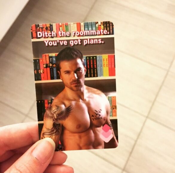 Amanda's hand holding a RT hotel key card with a picture of a shirtless, muscly tattooed dude standing in front of a bookshelf of Avon books