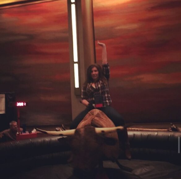 A blurry shot of amanda on a mechanical bull. she's wearing a plaid shirt and jeans, and has one arm raised 