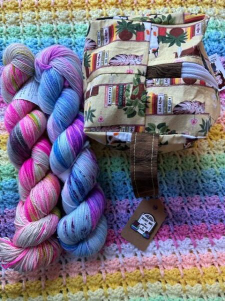A box shaped project bag next to two skeins of yarn. A pin below that. All of this is is staged on a rainbow crochet afghan.