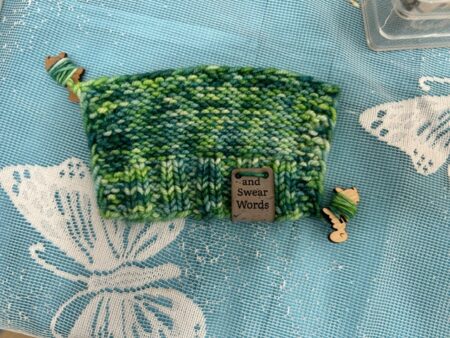 A green swatch of yarn with two sheep shaped tail minders wrapped up in the loose end and working end. A tag says And Swear Words