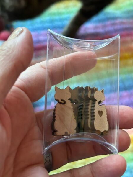 My hand holding a clear pouch of cat shaped tail minders. My actual cats leg and head in the background.