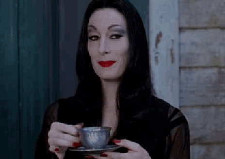 Morticia Addams lifts a cup of tea and smiles over the rim