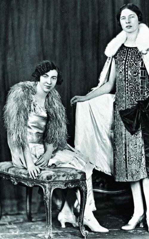 Ida cook, wearing a wavy bob, a shiny gown, low heels and a furry stole. Mary is standing with one arm showing off a cape with a fur trim wearing a gown that looks to be covered in velvet filigree. They look fabulous.