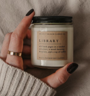 A small glass jar candle with a black lid being held by a hand with dark nails and a beige sweater cuff. The label says LIBRARY 