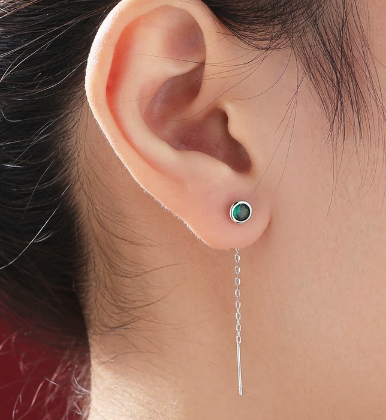 An ear with a green bezel set stone and a long silver chain behind the ear. 