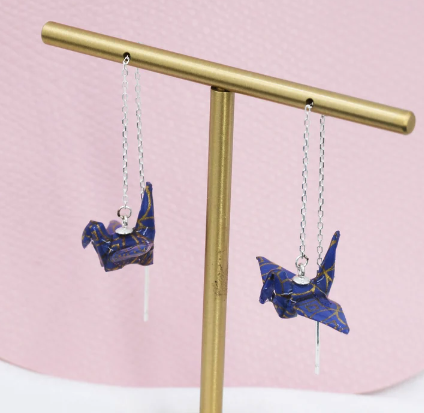 Miniature Blue origami cranes hang from a thin silver chain meant to thread through the ear. They're hanging from a gold colored T Bar 