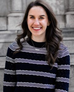 Sophie Vershbow is a white woman with long brown hair. She has blue eyes and is smiling. She is wearing a black sweater with white and purple horizontal stripes