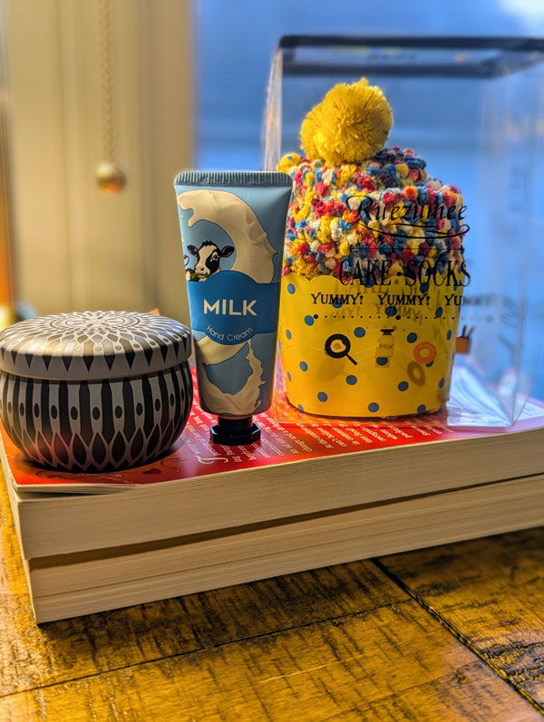 A small printed tin with a candle, a tube of MILK handcream, and a pair of fuzzy socks wrapped to look like a cupcake, on top of some books on my desk.