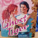 Ghost Tiger by someone named Carter. the sticker is over the name. Janice! Janice Carter. Thanks, Google. Anyway, there's a woman with curly hair in a ponytail and I think her hair is full on fuchsia. She's standing facing the side with her hand resting on the shoulder of a man facing the reader. He's looking off to the side, but his MULLET is Excellent. There is gold leaf printing of maybe daffodils up the side, and in the background is a pink washed image of a snarling tiger.