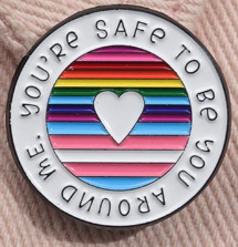 An enamel pin with a rainbow and trans and bi colors in stripes in the middle with a heart. At the border it says You're Safe to be You Around Me