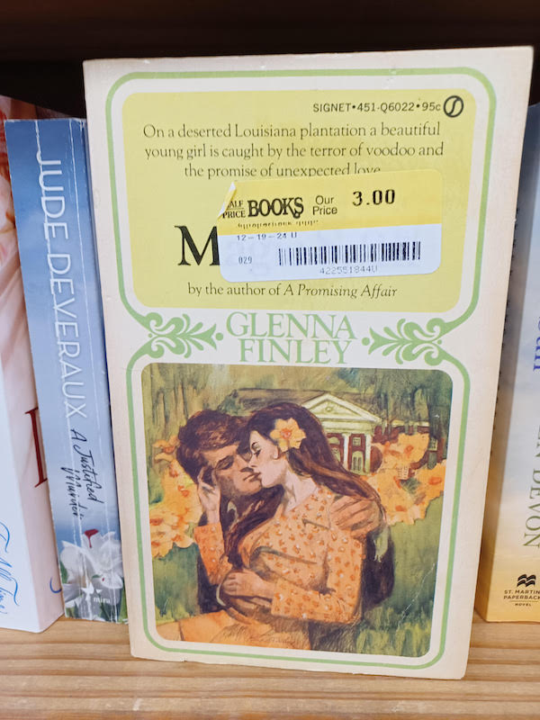 Love's Magic Spell by Glenna Finley, except the title is covered up by a price sticker. The top almost half is the author and title, and then an illustration of a man and woman in the grass in front of a large manor with columns. The man has brown hair and is mostly the behind the other character. She has a big daffodil in her hair, and is wearing an orange printed blouse with big collar points - MASSIVE. It's very 70s. 