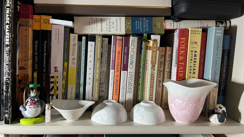 Three white teacups and a pink handle-less teapot with no lid lined up in a row on a shelf in front of tightly packed books. One of the white teacup’s mouth is flared in a leaf-like shape, while the other two are perfect bowl shapes. The teapot is a blush pink gradient that becomes white at the top. All the ceramics have holographic like splotches, giving them a mother of pearl sort of look in places. To the left side of the shelf, next to the leaf shaped teacup, is a polymer clay figurine of Totoro with a red bow tie, a baby Totoro on their head, and a leaf umbrella they’re holding over both their heads. There is a tiny corked glass jar filled with very small pieces of rose quartz between Totoro and the teacup. To the right side of the shelf, next to the teapot, there is a figurine of a panda on their back. Their name is Pandan Rollsalot.