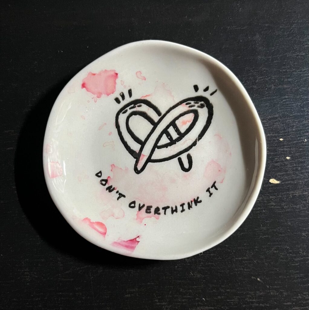 A small, round sauce plate on a black desk. It has an adorable pretzel in the middle and the words, “Don’t overthink it” written in an arc and all caps under the pretzel. There is a bit of dried red ink on the sauce plate.