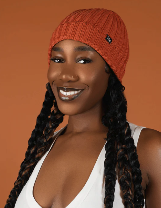 A Black woman with long braids smiles at the camera. They are wearing a orange rib knit beanie on their head, with braids emerging from beneath the hem